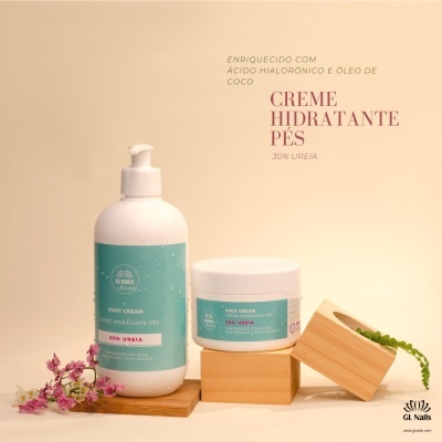 Foto de dois cremes hidratantes para pés em embalagem branca com rótulo azul-turquesa, sobre base de madeira com flores e planta ao fundo.