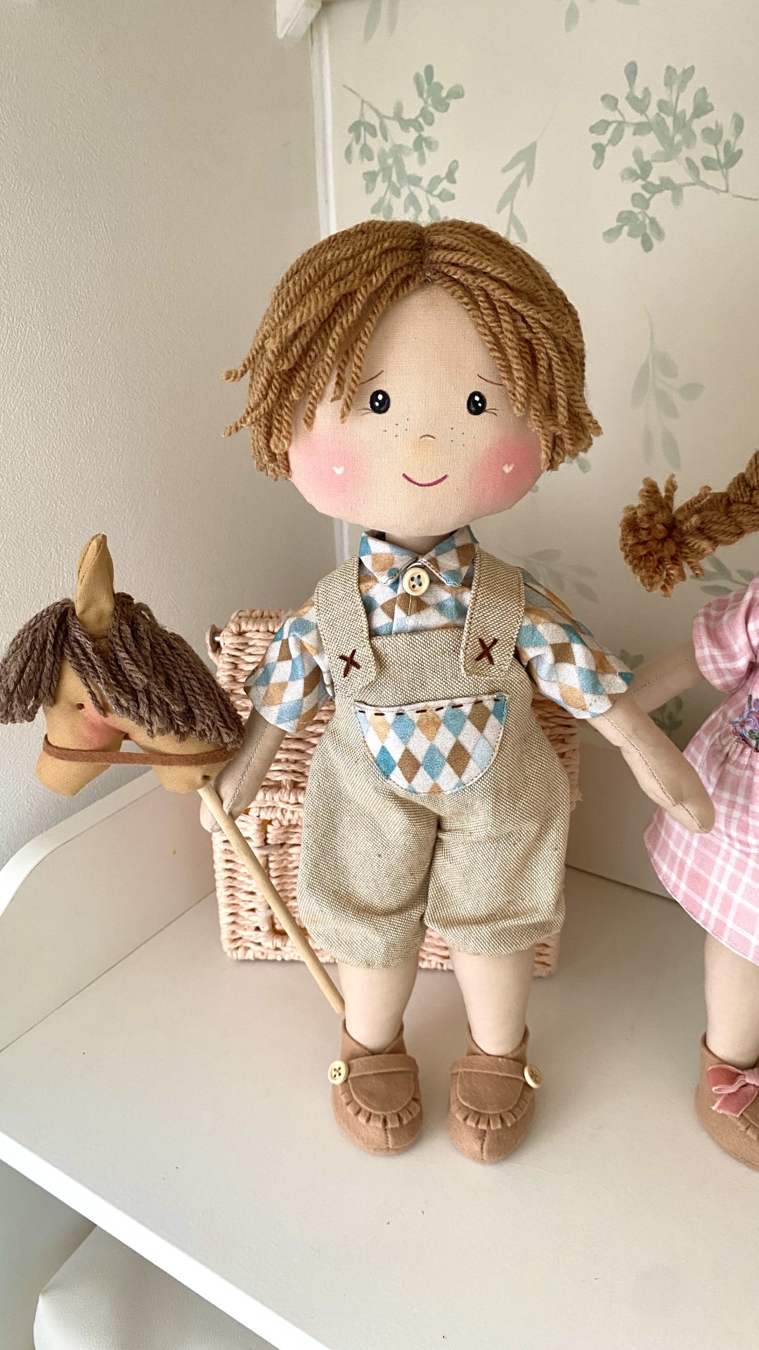 Kit de costura boneco Pedro Boneco de pano com roupa bege e azul segurando cavalo de pau ao lado de cesta e papel de parede floral