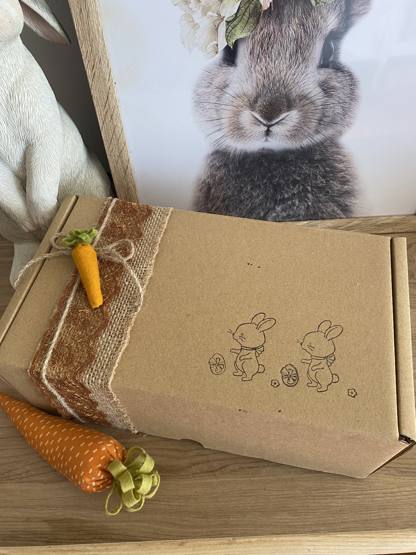 Mistery box especial Páscoa 2024 Caixa de cartão castanho decorada com fita rústica e miniatura de cenoura, com desenho de coelhos, sobre mesa de madeira