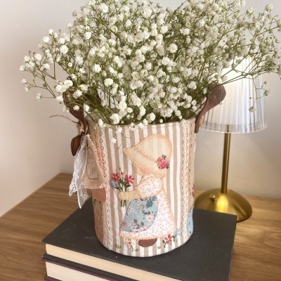 Vaso decorativo de tecido com boneca e flores brancas em cima de livros numa mesa