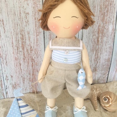 Boneco de pano com roupa azul e bege e cabelo castanho, segurando peixe de tecido azul. Barquinho de madeira e conchas ao lado.