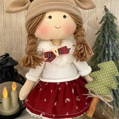 Boneca decorativa com chifres e roupa festiva, segurando árvore verde, com lanterninha e árvore decorativa ao fundo