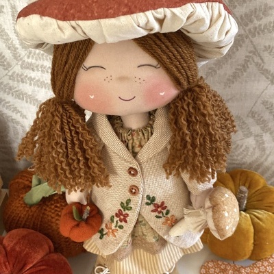Boneca de pano com chapéu de cogumelo e vestuário bordado, rodeada de abóboras decorativas em tecido