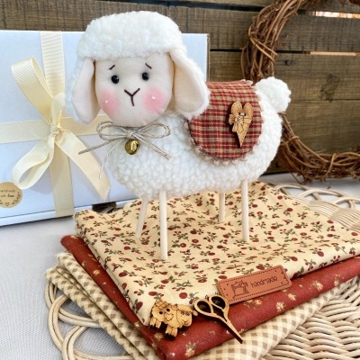 Ovelha decorativa branca com manta xadrez vermelha em tecidos florais e xadrez sobre superfície de verga