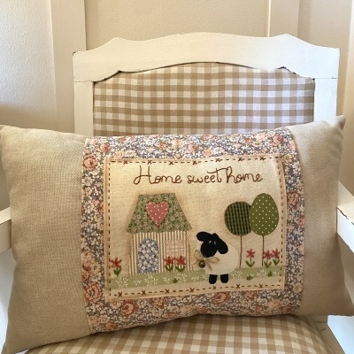 Almofada bege com aplicação colorida e bordado Home sweet Home em cadeira branca com almofadão xadrez