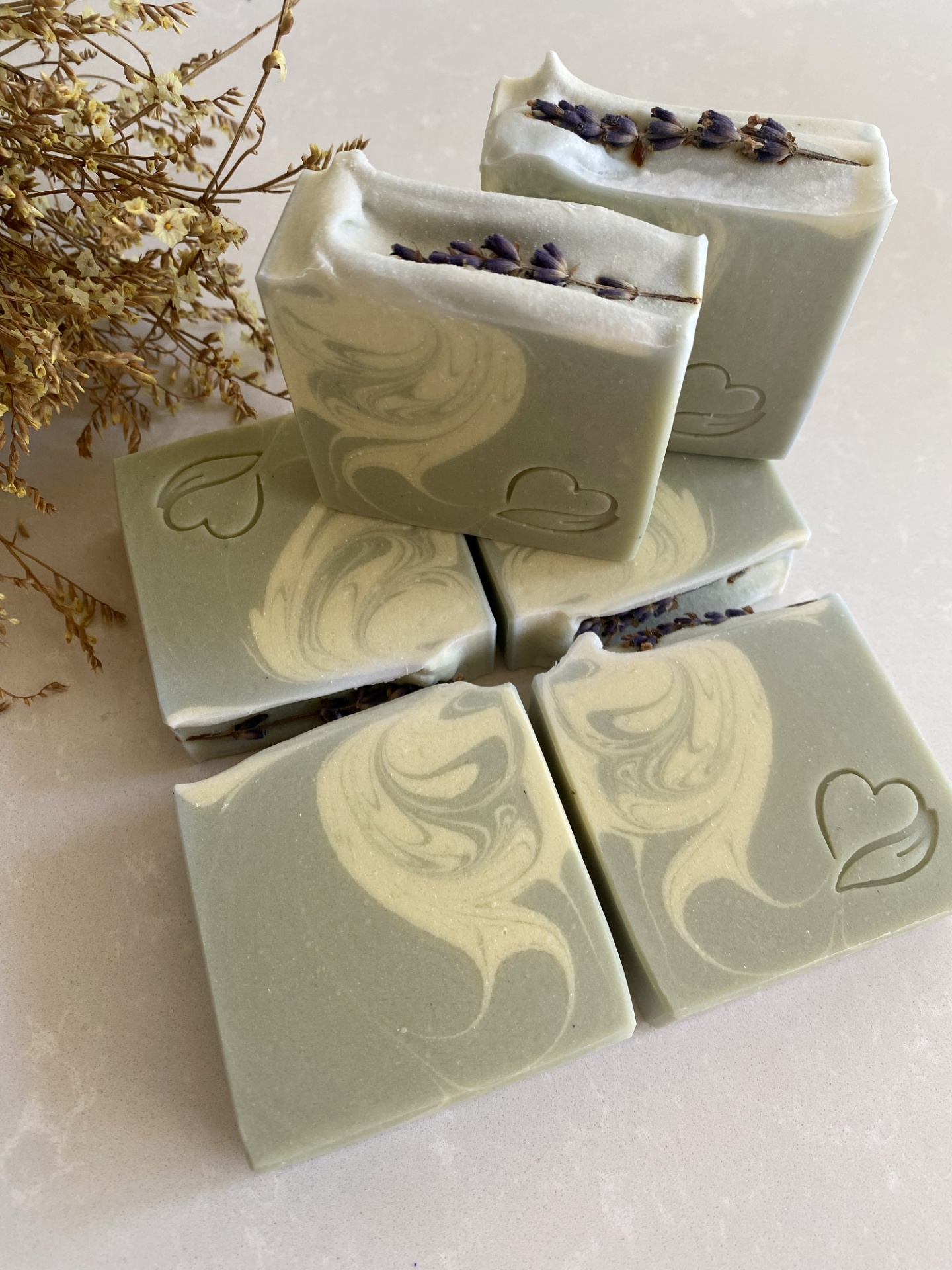 sabonete natural de lavanda e alecrim Seis sabonetes verdes com padrão creme e flores secas, sobre superfície clara com coração gravado.