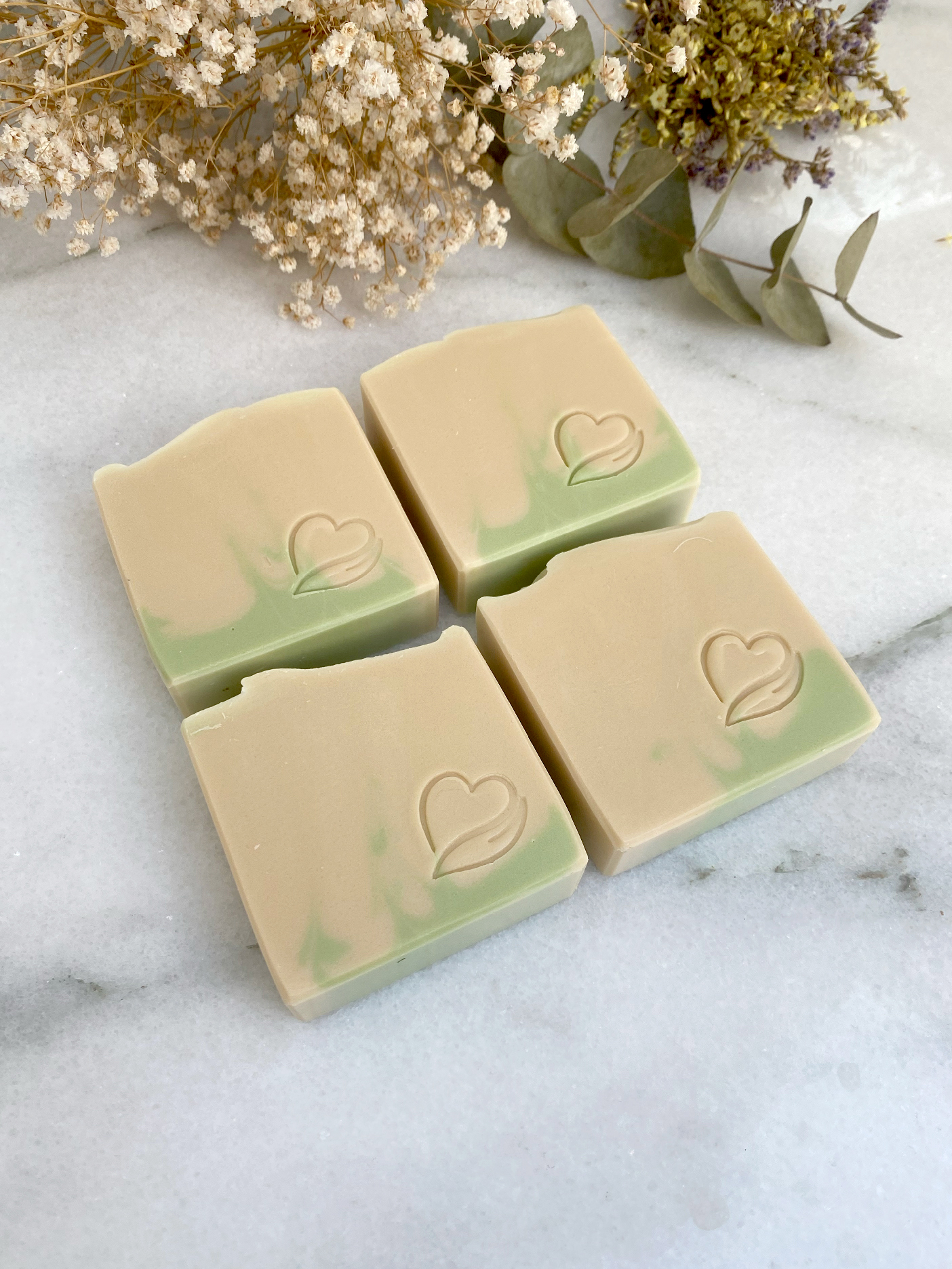 Sabonete Natural erva príncipe Quatro barras de sabão bege e verde com um coração gravado, sobre mármore branco e flores secas ao fundo