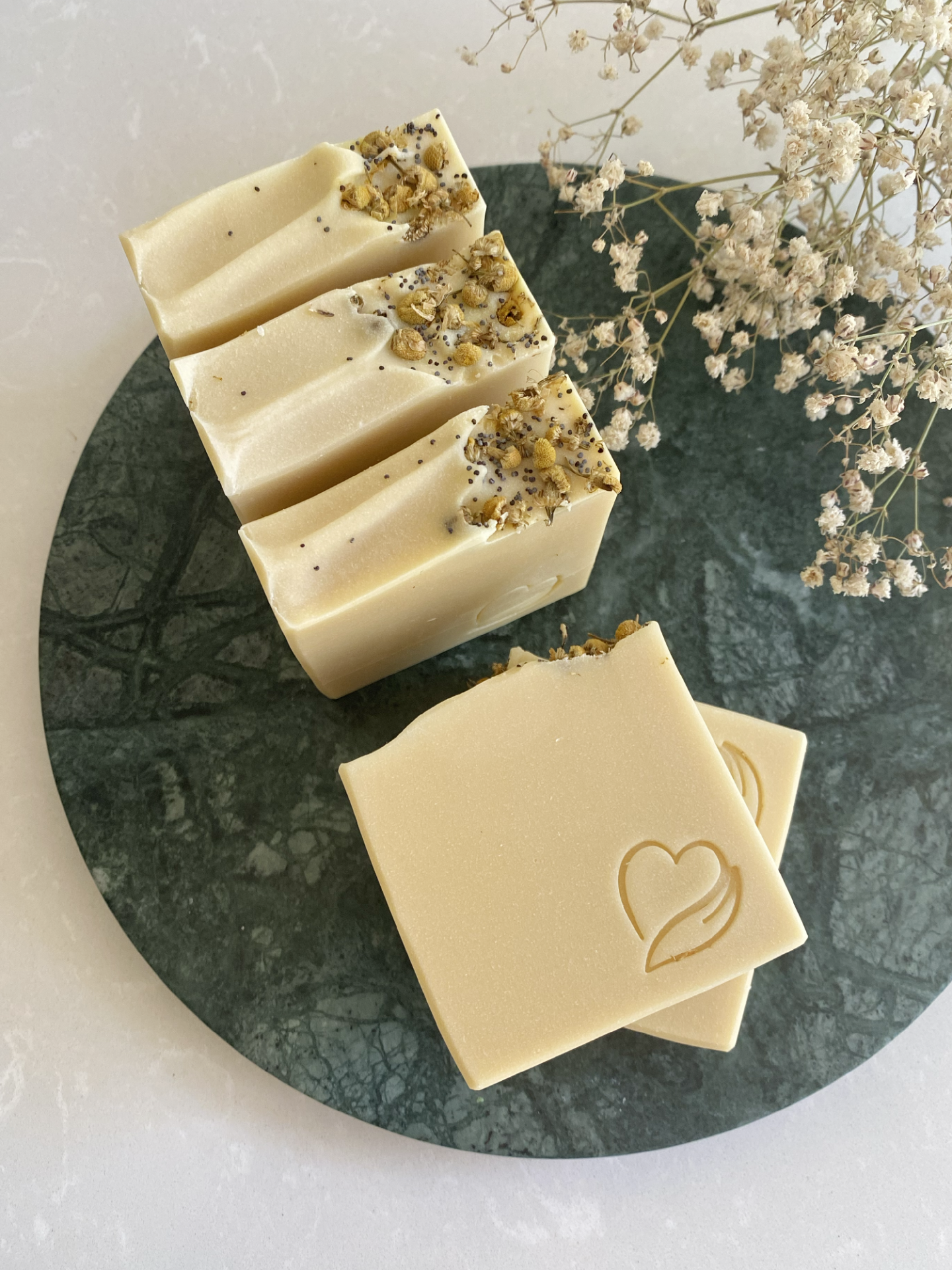 Sabonete Natural Leite de cabra Sabonetes sólidos creme com flores secas e sementes decorativas, em prato verde, junto a flores secas brancas.