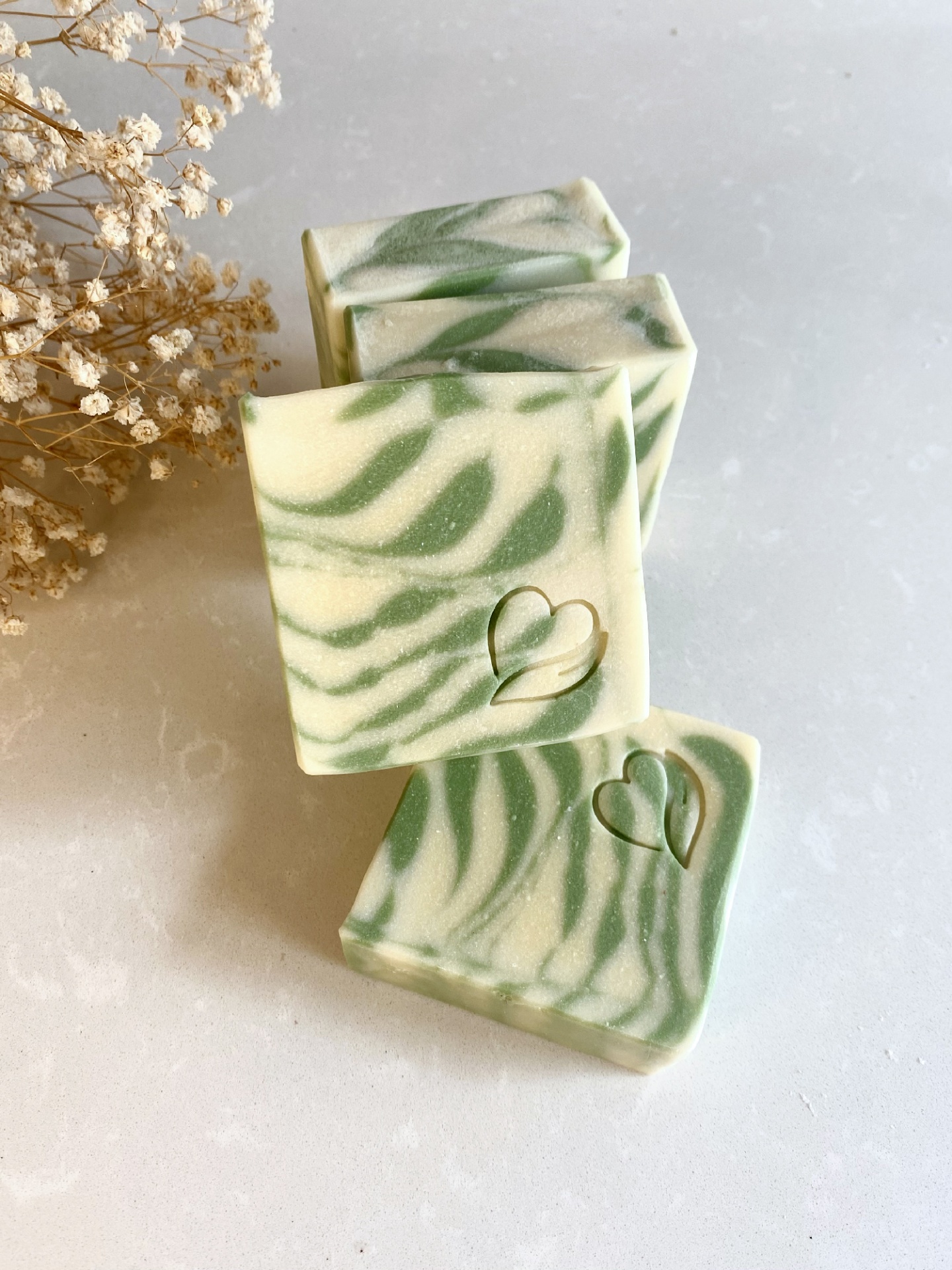 Sabonete Natural de Limão Sabonetes verde e branco com padrão ondulado, decorados com coração gravado, ao lado de flores secas.