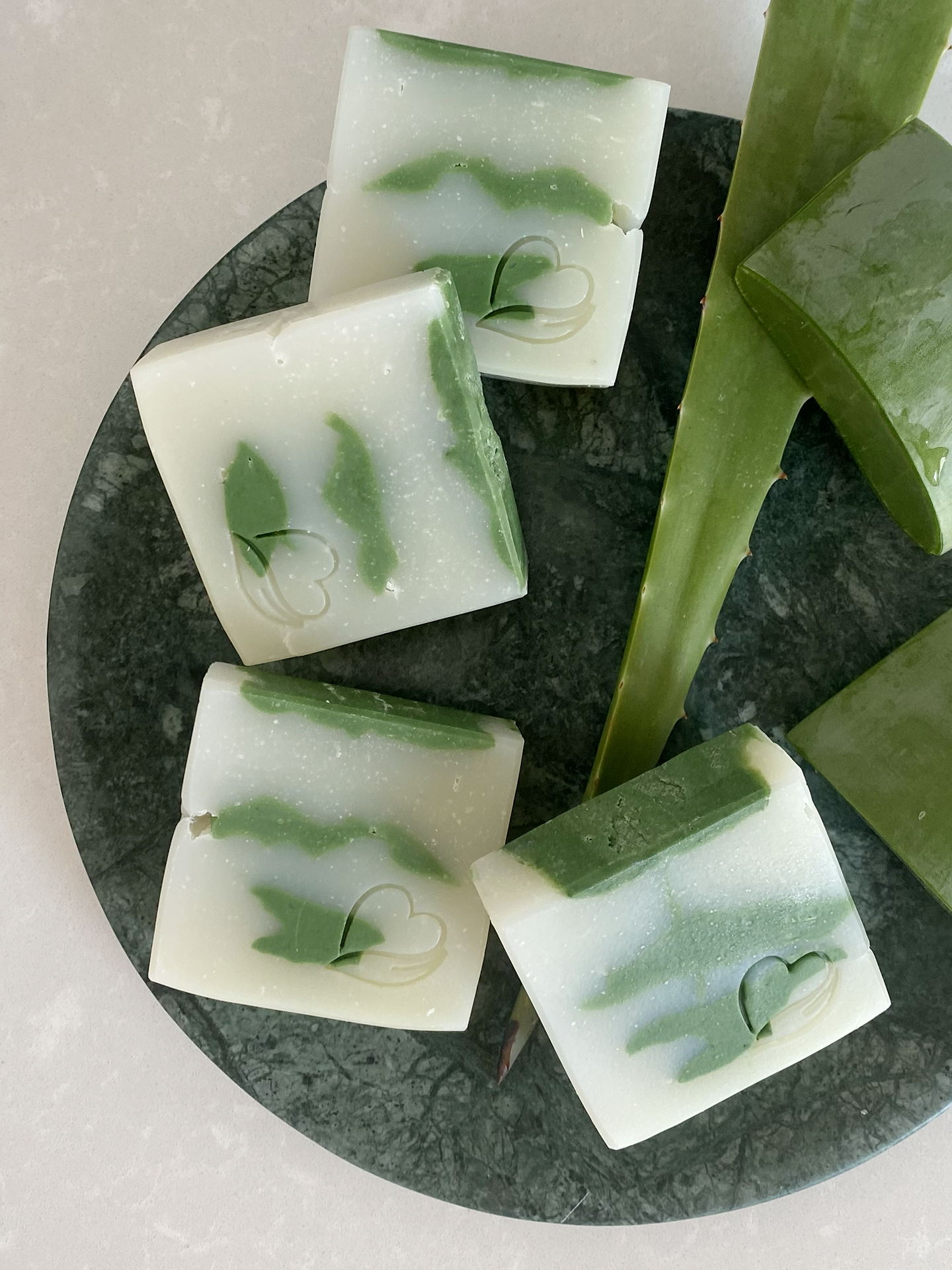 Sabonete Natural de Aloé Vera Sabonetes quadrados verdes e brancos com folhas de aloe vera ao lado e prato escuro.