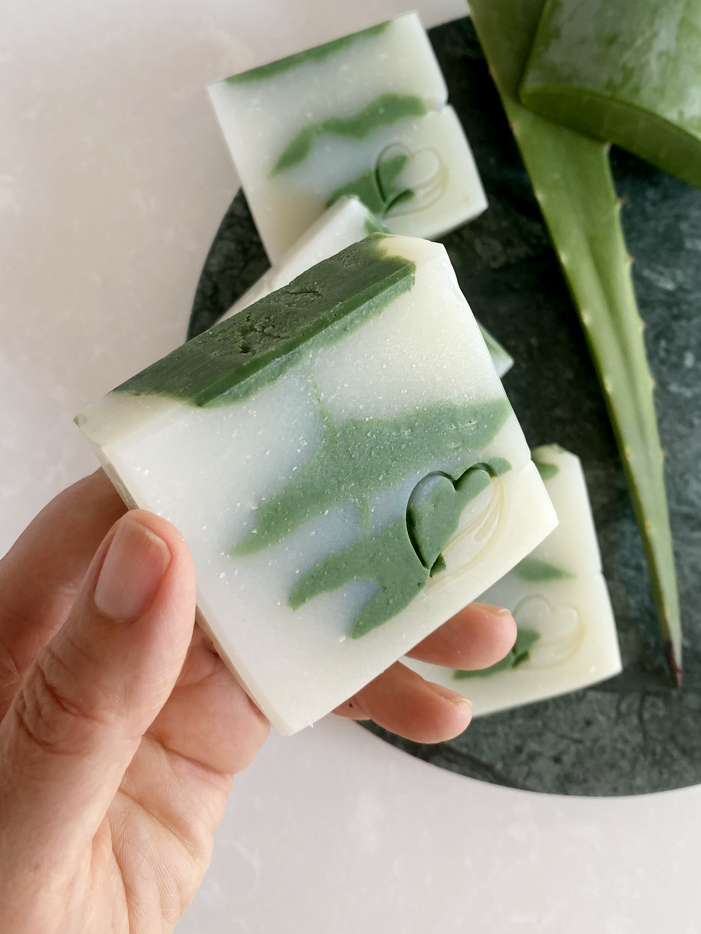 Sabonete Natural de Aloé Vera Sabonetes brancos e verdes marmoreados com relevo em coração e folha de aloe vera ao fundo