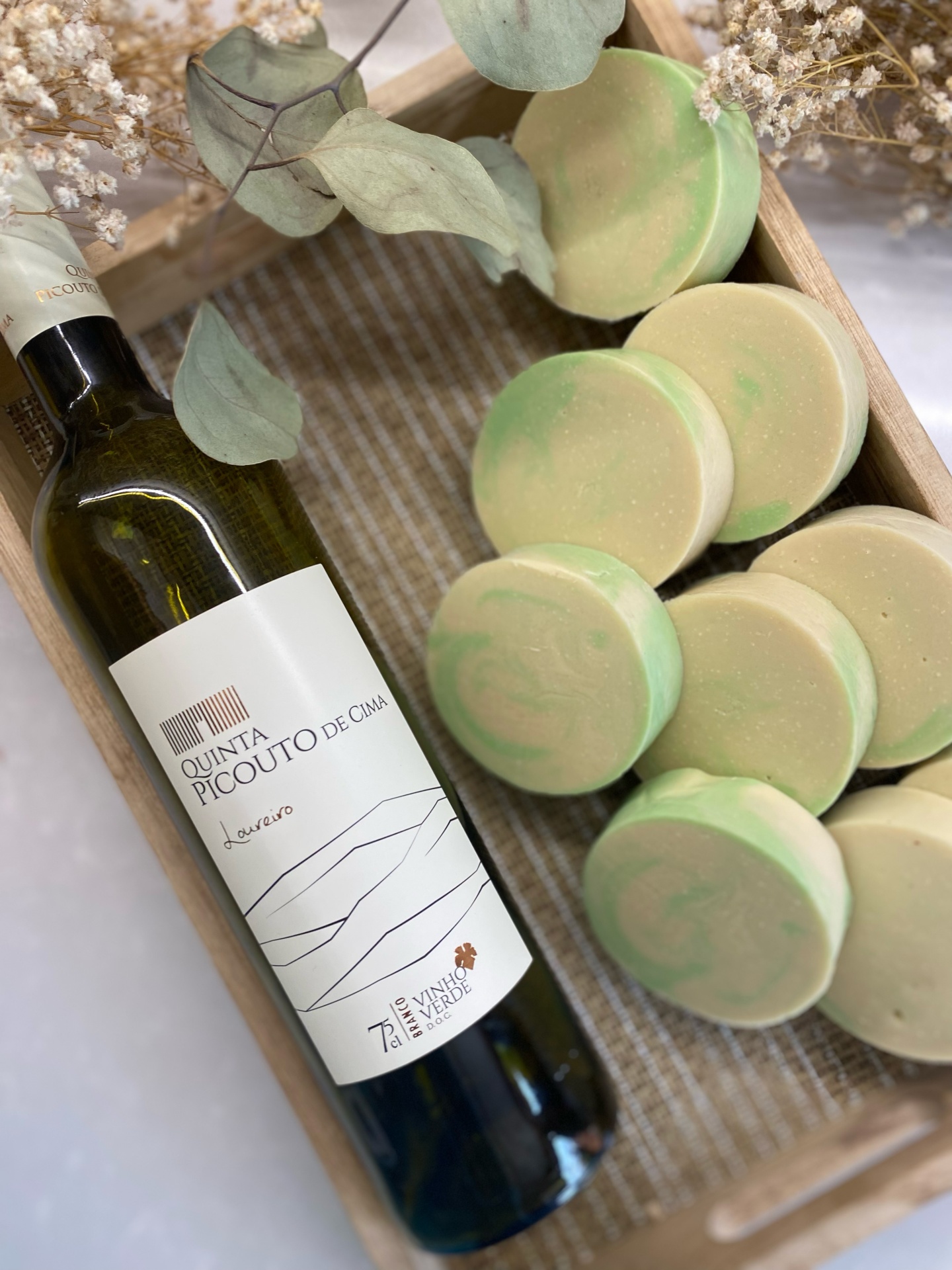 Workshop Sabonetes Naturais de Vinho Garrafa de vinho branco e sabonetes redondos verdes numa caixa de madeira com folhas secas