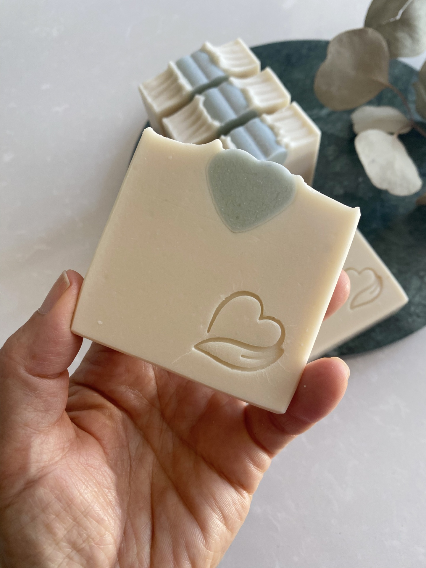 Sabonete Natural Coco Fresh Sabonete quadrado bege com detalhe de coração verde e gravação em relevo, segurado por uma mão.