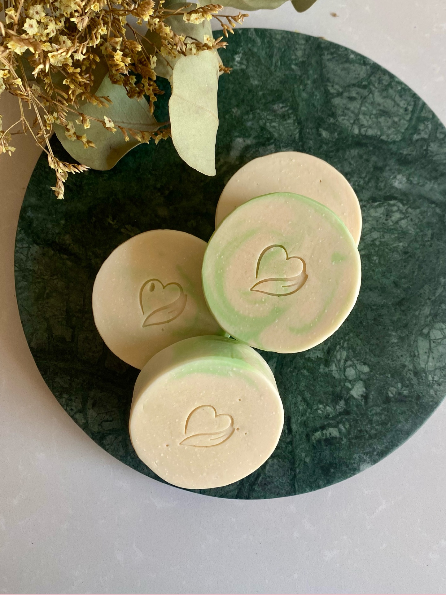 Sabonete natural de vinho branco Sabonetes redondos creme e verde com logo em relevo num prato de mármore verde e flores secas