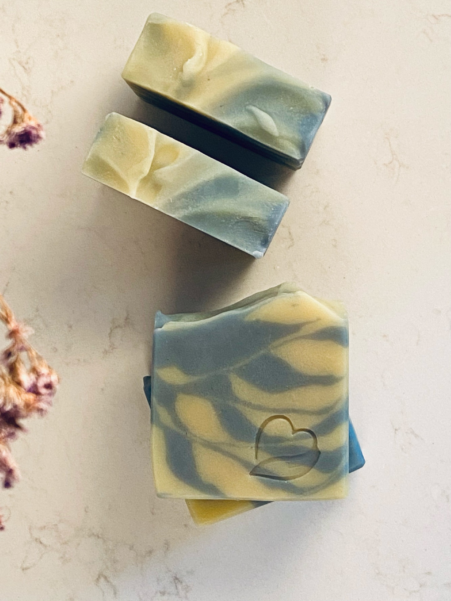Sabonete Natural Alecrim e Menta 2 em 1 Três sabonetes artesanais com padrão marmoreado amarelo e azul sobre superfície clara