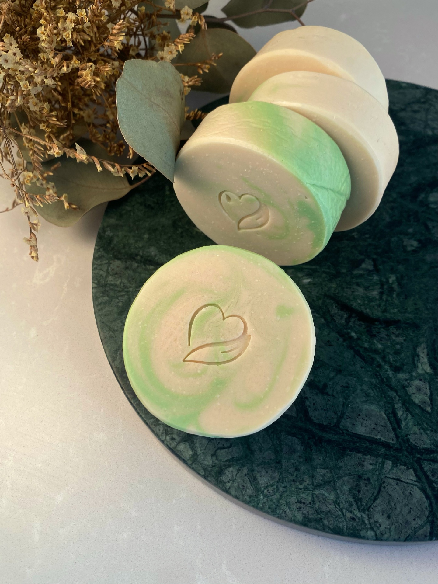 Sabonete natural de vinho branco Sabonetes redondos bege e verde claro com símbolo de coração gravado, sobre prato de pedra verde escura com folhas secas ao fundo