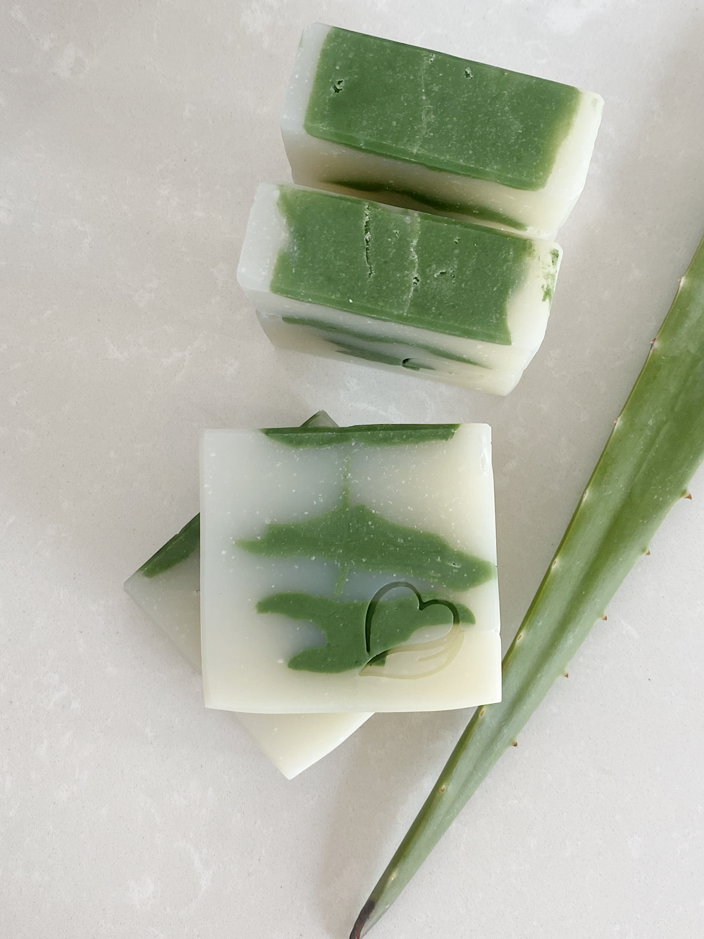 Sabonete Natural de Aloé Vera Barras de sabão artesanal verde e branco com relevo em formato de coração e folha de aloé-vera ao lado