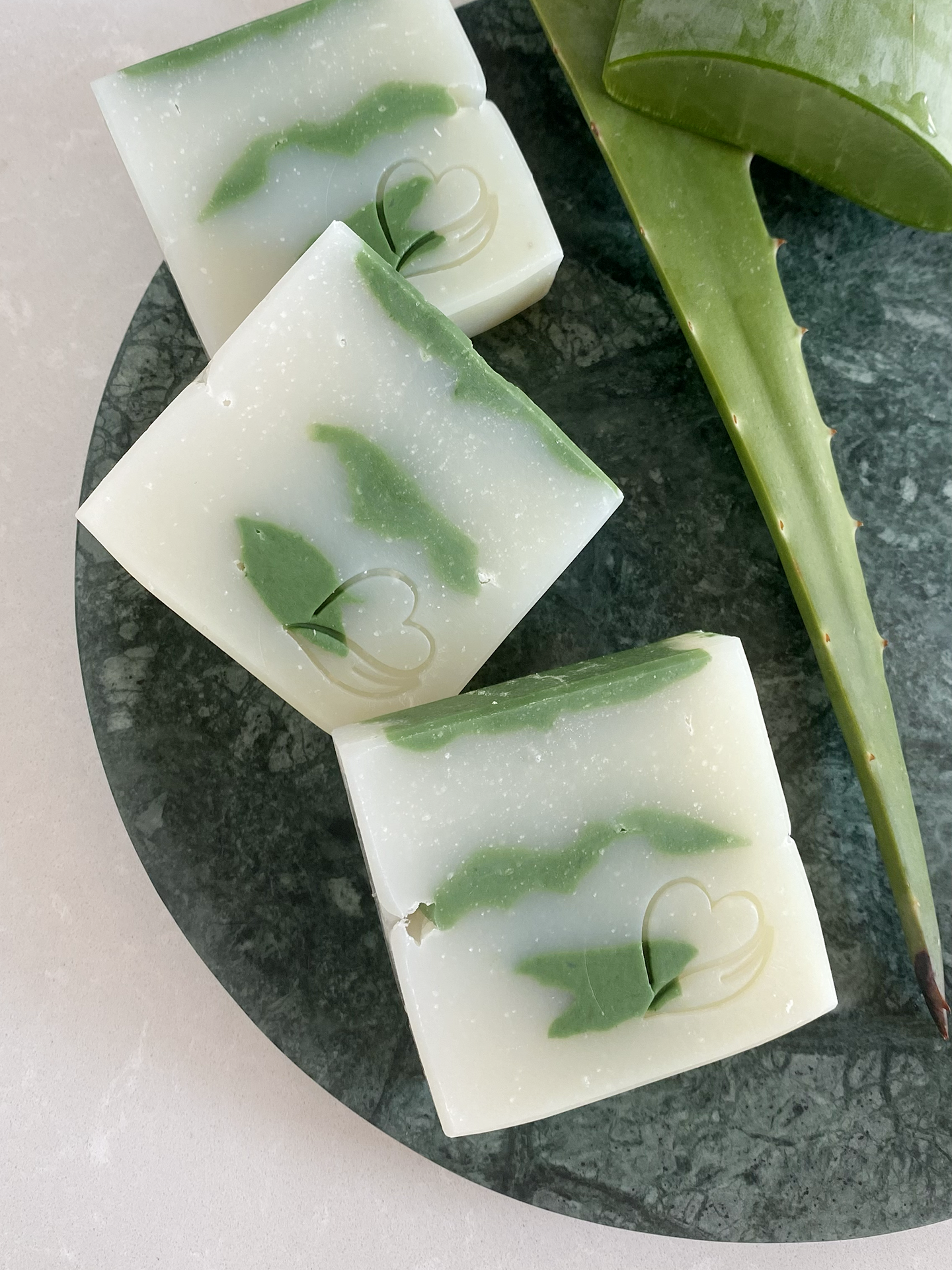 Sabonete Natural de Aloé Vera Barras de sabão branco e verde com gravação de coração sobre prato de pedra e folha de aloe vera.