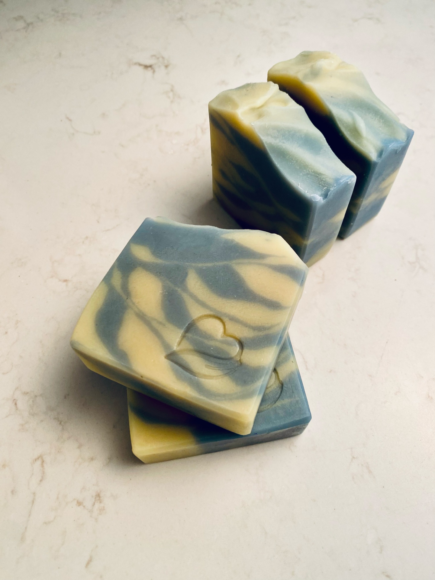 Sabonete Natural Alecrim e Menta 2 em 1 Quatro sabonetes quadrados azul e bege com padrão marmoreado e selo de coração com folha.