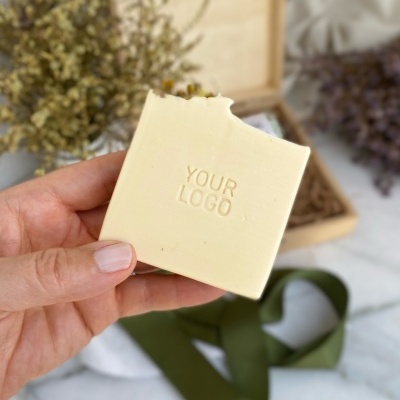 Sabonete quadrado creme com inscrição YOUR LOGO segurado por mão contra fundo com caixa, fitas verdes e flores secas