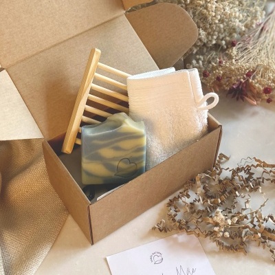 Caixa aberta com sabonete marmoreado, saboneteira de madeira, toalha branca e cartão obrigada Mãe com flores secas