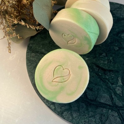 Sabonetes redondos bege e verde claro com símbolo de coração gravado, sobre prato de pedra verde escura com folhas secas ao fundo
