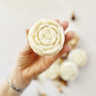 Mão segurando sabonete branco em forma de flor com outros sabonetes ao fundo