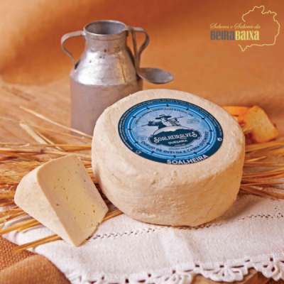 Queijo redondo de ovelha com rótulo azul, fatia de queijo, jarra metálica e toalha branca rendada.