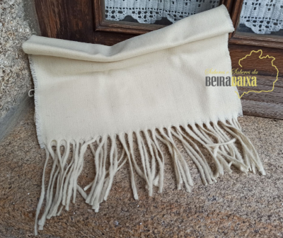 Echarpe creme com franjas sobre superfície de pedra e madeira com janela rendada ao fundo