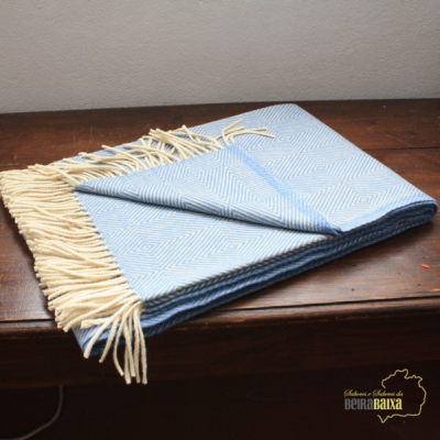 Manta azul com padrão geométrico branco e franjas bege sobre mesa de madeira escura