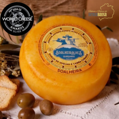 Queijo redondo amarelo com rótulo Soalheira Alves, azeitonas e pão sobre toalha branca