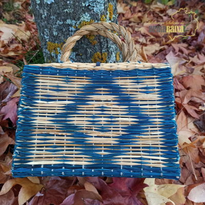 Mala de palha trançada azul e bege com asa trançada natural em ambiente de folhas secas