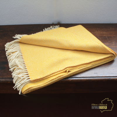 Manta amarela com padrão geométrico e franjas brancas sobre mesa de madeira