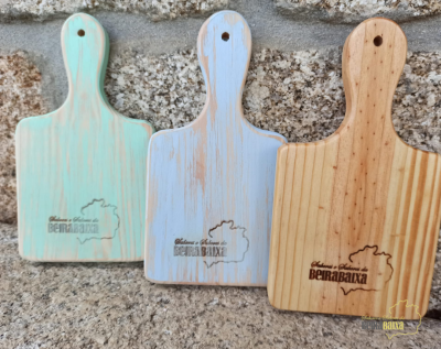 Três tábuas de corte de madeira com cabos em azul claro, azul esbatido e natural, com texto BERABAXA, sobre pedra rústica