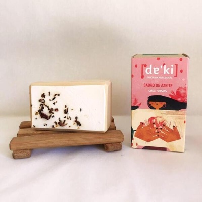 Sabonete branco com pedaços escuros em saboneteira de madeira ao lado de caixa rosa de sabonete artesanal de azeite com rosas