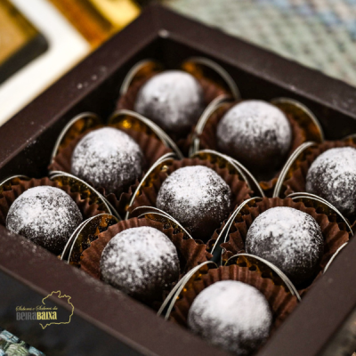 Caixa preta com bombons de chocolate polvilhados com açúcar em pó em forminhas castanhas, com logótipo na tampa.