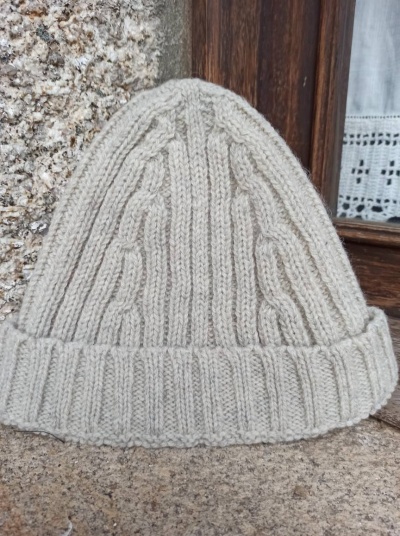 Gorro de malha bege com padrão de trança colocado sobre pedra em fundo de madeira e cortina branca.