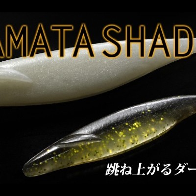 Dois iscos de pesca SAKAMATA SHAD, branco e preto com brilhos dourados, em fundo preto.