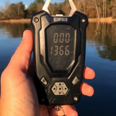 Dispositivo digital preto Rapala com visor mostrando números, segurado à beira de um lago