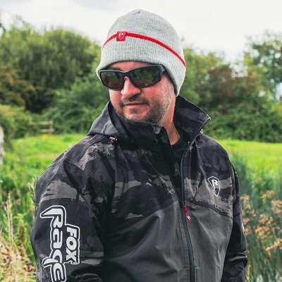 Homem com casaco Fox Rage preto e boné cinzento numa zona verde junto a água