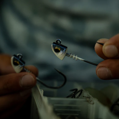 Gancho de pesca com cabeça metálica prateada e amarela segurado por mãos próximas a uma caixa de ganchos