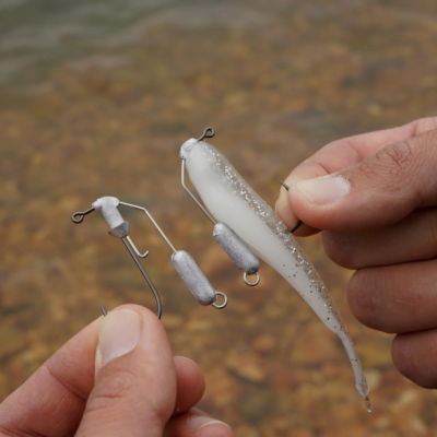 Iscos artificiais de pesca brancos e prateados nas mãos, com ganchos e pesos, sobre água e pedras.
