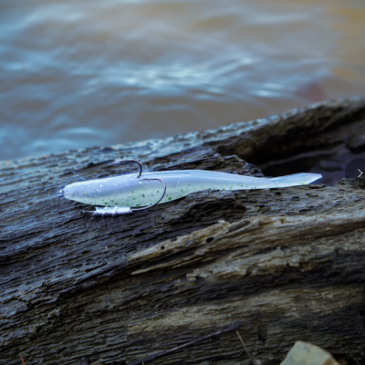 Isca artificial prateada com gancho de pesca sobre um tronco à beira da água.