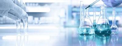Frascos de laboratório com líquidos coloridos e mãos com luvas azuis manipulando tubos de ensaio