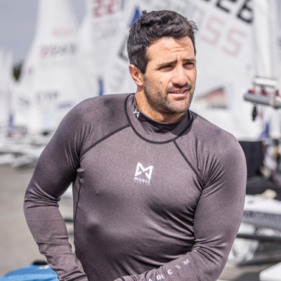 Homem com camisa preta justa com logotipo M@BASICS e veleiros ao fundo