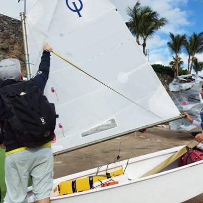 Barco de vela pequeno branco com vela branca e símbolo azul, pessoas a manipular a vela junto a cais com palmeiras.