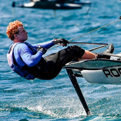 Homem com roupa desportiva azul e preta a velejar num barco hidrofoil