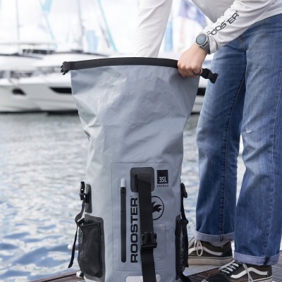 saco impermeável cinzento claro com logótipo ROOSTER num cais junto a água e barcos