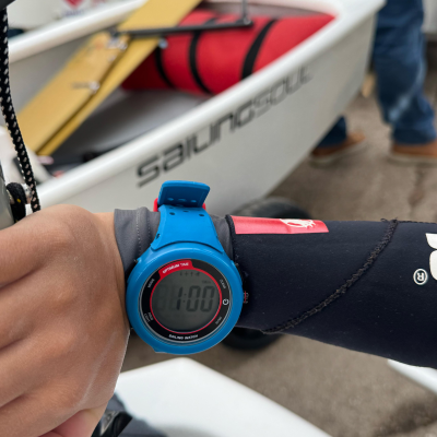 Relógio digital azul num pulso com fato de neopreno negro ao lado de embarcação branca e prancha amarela