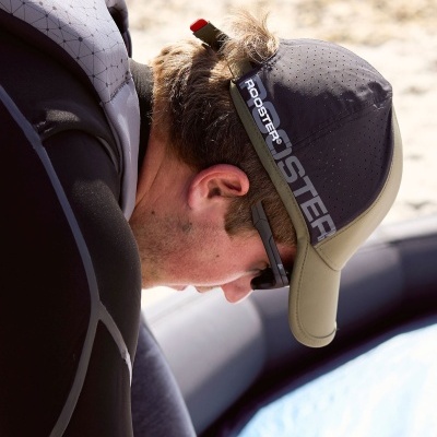 Pessoa de boné cinza escuro e bege com texto ROOSTER, óculos escuros e roupa de neopreno preta junto a superfície azul.